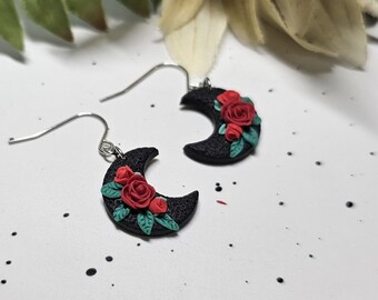 May include: A pair of drop earrings featuring black crescent moon charms with a textured surface. Each moon is decorated with small red roses and green leaves. The charms are attached to silver-toned metal earring hooks for pierced ears.