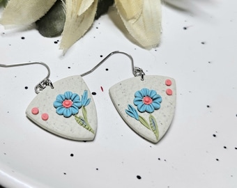 May include: A pair of triangular-shaped earrings featuring a speckled off-white base with a raised blue flower design, a pink centre, and green leaves. The earrings are attached to silver-toned metal hooks and include small pink circular accents.