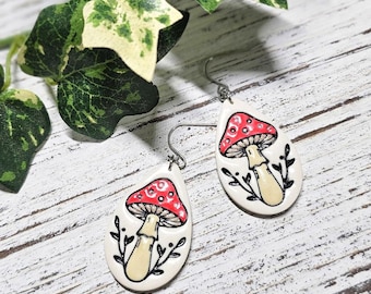 May include: A pair of teardrop-shaped earrings featuring a hand-painted red and white mushroom design on a cream background. Each earring includes a black ink illustration of small leafy branches at the base and a silver-toned metal hook.