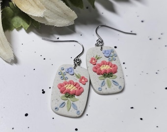 May include: A pair of rectangular drop earrings featuring a speckled light grey base with a raised floral design. The flowers are coloured in shades of pink, yellow, and light blue, accented with green leaves and stems, attached to silver-toned metal hooks.