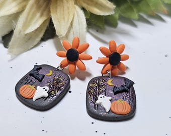 May include: A pair of dangle earrings featuring orange flower studs with black centres. Each earring has a square dark purple pendant decorated with a white ghost, an orange pumpkin, a black bat, a yellow crescent moon, and dark tree silhouettes.