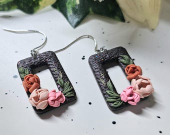 May include: A pair of rectangular dangle earrings featuring a textured black base with subtle glitter. Each earring is decorated with small, sculpted clay flowers in shades of terracotta, blush, and light pink, accented by tiny green leaves.