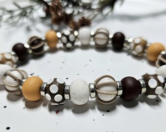 May include: A beaded bracelet featuring a repeating pattern of round and ribbed beads in shades of white, tan, and dark brown. The beads include solid colours, speckled textures, and white polka dot patterns, separated by small, reflective silver-toned spacers.
