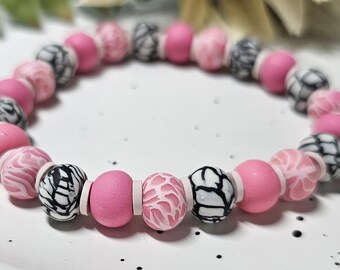 May include: A circular beaded bracelet featuring alternating round beads in solid matte pink, white and pink marbled patterns, and white beads with black crackle-like veining, separated by small, flat, light pink disc-shaped spacer beads.