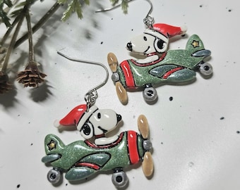May include: A pair of drop earrings featuring a white cartoon dog wearing a red Santa hat, piloting a glittery green aeroplane with red accents, a tan propeller, and silver-toned metal hooks for attachment to the earlobes.