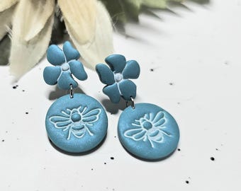 May include: A pair of drop earrings featuring a matte blue flower stud connected to a circular blue pendant. Each circular pendant is embossed with a white bee design. The earrings are crafted from a smooth material and hang from silver-toned metal jump rings.
