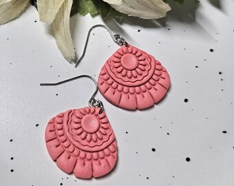 May include: A pair of coral pink drop earrings featuring a semi-circular fan shape with a raised, embossed radial pattern. The earrings are attached to silver-toned metal fishhook wires, showcasing a textured, matte finish on the surface.