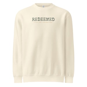 May include: Cream-colored sweatshirt with the word "REDEEMED" printed in dark green, with the phrase "(That was unexpected)" below it. The long-sleeved crew neck top is made of a soft material.