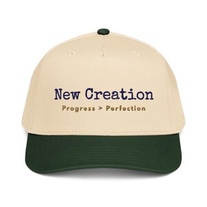 May include: A cream-colored baseball cap with a dark green brim and button. The cap features the text "New Creation" in navy blue, with "Progress > Perfection" in a smaller, gold font below.