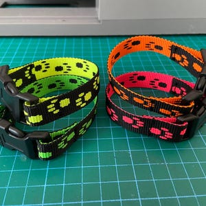 Puede incluir: Dos collares para mascotas con hebillas de plástico negro. Uno es verde neón con huellas de patas negras, y el otro es naranja y rosa con huellas de patas negras. Los collares están sobre una superficie de cuadrícula verde.
