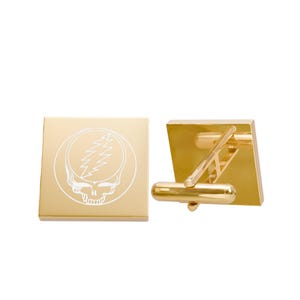 May include: Gold-toned cufflinks with a square face featuring a white skull and lightning bolt design within a circle. The cufflinks have a classic post and toggle closure. The design is a stylized skull with a lightning bolt.
