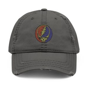May include: Distressed gray baseball cap featuring a colorful emblem with a skull and lightning bolt design. The emblem is in a circle with red, blue, and yellow colors. The cap has a pre-worn, vintage look.