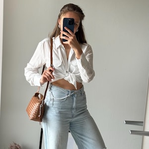 May include: A white button-down shirt tied at the waist, paired with light-wash denim jeans featuring a distressed knee. A brown woven crossbody bag hangs from the shoulder. The outfit is completed with a pearl chain belt.
