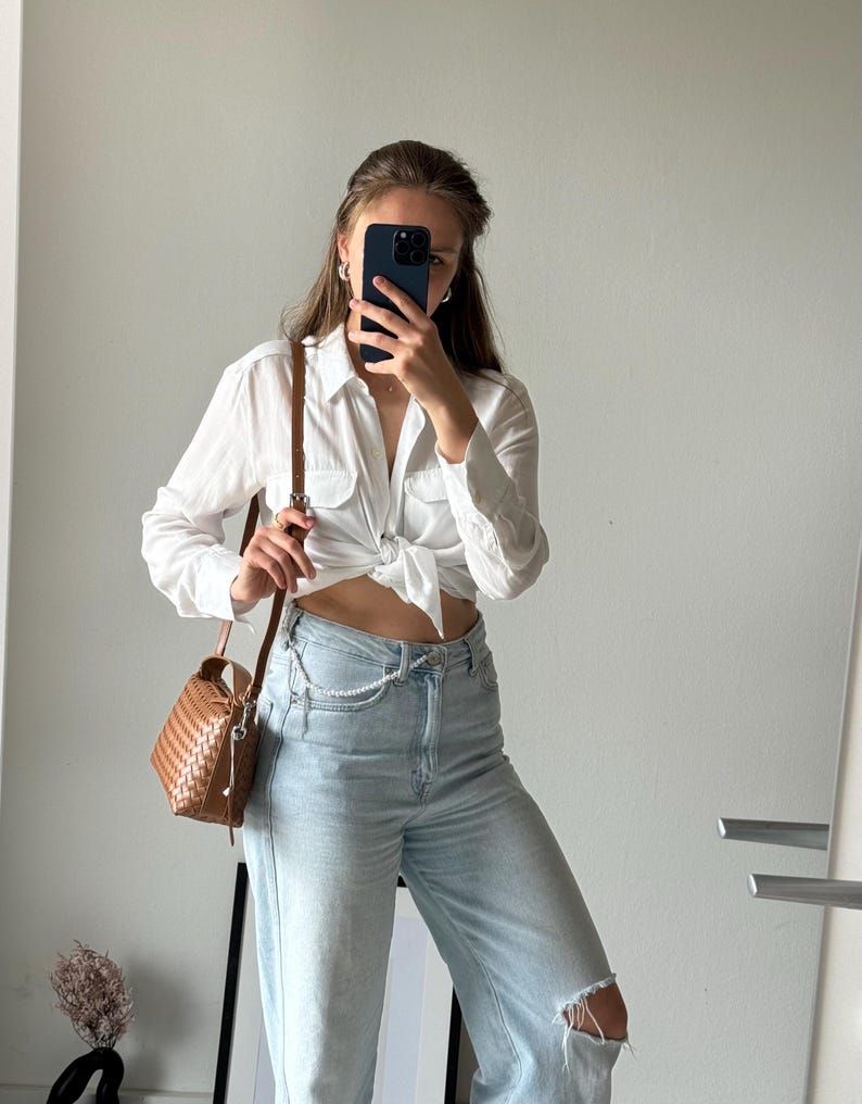 May include: A white button-down shirt tied at the waist, paired with light-wash denim jeans featuring a distressed knee. A brown woven crossbody bag hangs from the shoulder. The outfit is completed with a pearl chain belt.