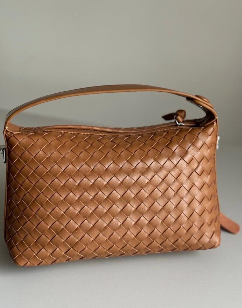 May include: A woven brown handbag with a top handle and a zipper closure. The bag has a rectangular shape and a textured surface. The handle is a matching brown color.