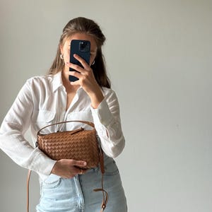 May include: A woven brown leather crossbody bag with a long strap. The bag is rectangular with a zipper closure and a top handle. The bag is held by a person wearing a white button-down shirt and light blue jeans.