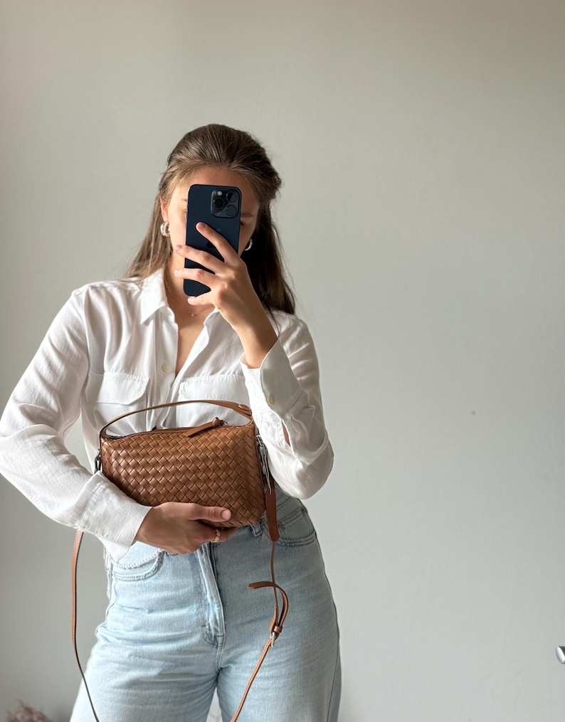 May include: A woven brown leather crossbody bag with a long strap. The bag is rectangular with a zipper closure and a top handle. The bag is held by a person wearing a white button-down shirt and light blue jeans.