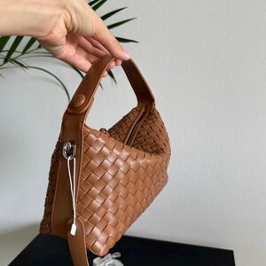 May include: A brown woven leather handbag with a top handle and a zipper closure. The bag has a tag attached with a silver clasp. The bag is held up against a white background.