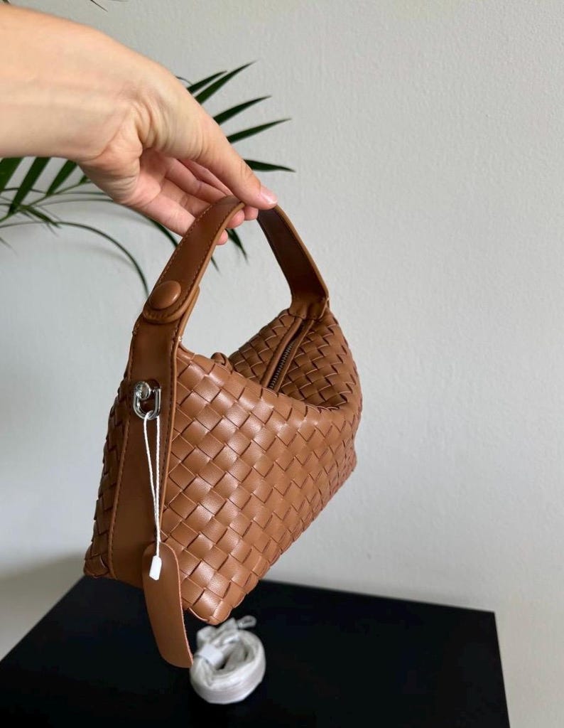 May include: A brown woven leather handbag with a top handle and a zipper closure. The bag has a tag attached with a silver clasp. The bag is held up against a white background.