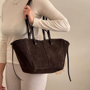 Könnte beinhalten: Dunkelbraune Wildleder-Tragetasche mit Ledergriffen und einem langen Schulterriemen. Die Tasche hat eine rechteckige Form mit einer breiten Öffnung und einem Knopfverschluss. Die Tasche wird von einer Person gehalten.