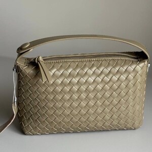 May include: A woven, rectangular handbag in a muted olive green hue. The bag features a top handle and a long, adjustable strap. The zipper closure is accented with a leather pull. The bag's design is characterized by its intricate woven pattern.
