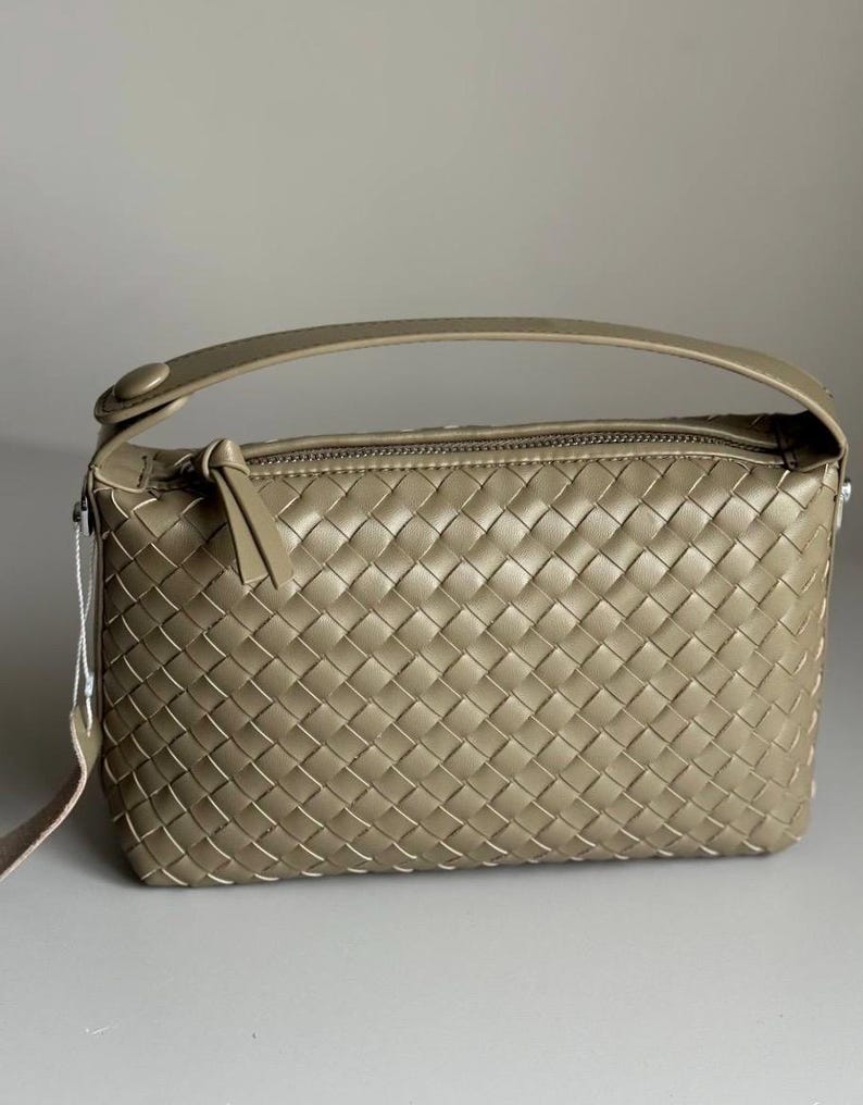 May include: A woven, rectangular handbag in a muted olive green hue. The bag features a top handle and a long, adjustable strap. The zipper closure is accented with a leather pull. The bag's design is characterized by its intricate woven pattern.
