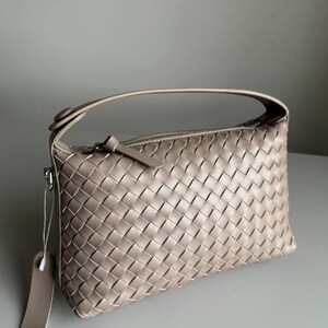 May include: A beige woven leather handbag with a top handle and a zipper closure. The bag has a rectangular shape and a tag attached to the side. The woven pattern is consistent throughout the bag's exterior.