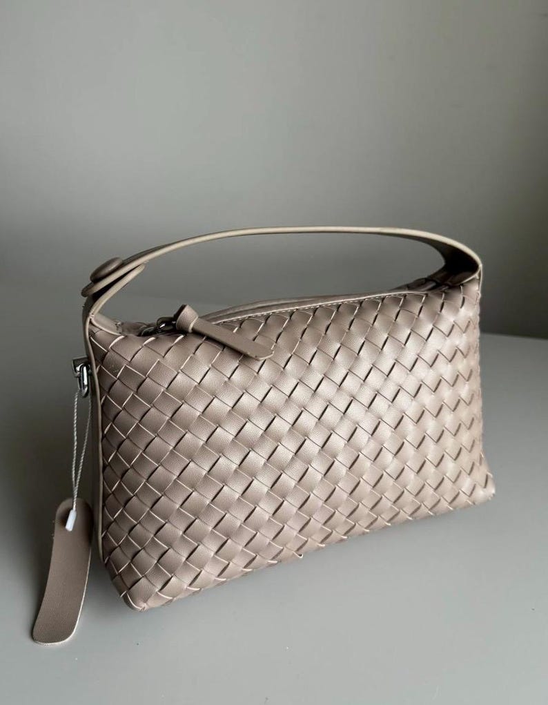 May include: A beige woven leather handbag with a top handle and a zipper closure. The bag has a rectangular shape and a tag attached to the side. The woven pattern is consistent throughout the bag's exterior.