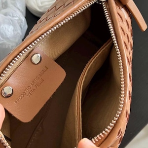 May include: A close-up of a brown leather handbag with a woven design. The bag has a silver zipper and a small leather tag inside with the text "PRODOTTO ARTIGIANALE VERA PELLE". The interior is a lighter brown color.