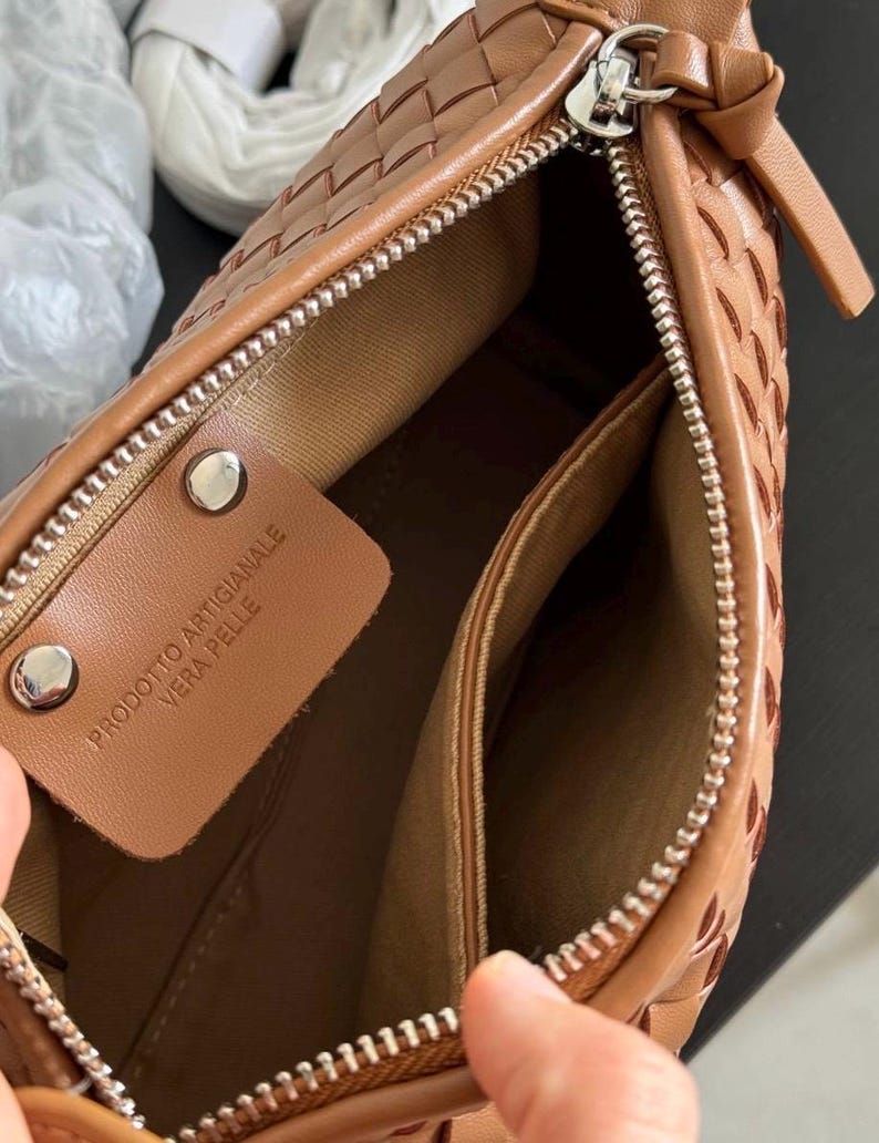 May include: A close-up of a brown leather handbag with a woven design. The bag has a silver zipper and a small leather tag inside with the text "PRODOTTO ARTIGIANALE VERA PELLE". The interior is a lighter brown color.