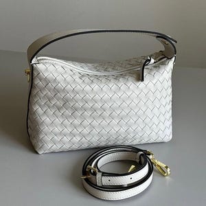 May include: White woven leather handbag with a top handle and a detachable shoulder strap. The bag features a zipper closure and gold-tone hardware. The strap is white with black trim and gold-tone clasps.