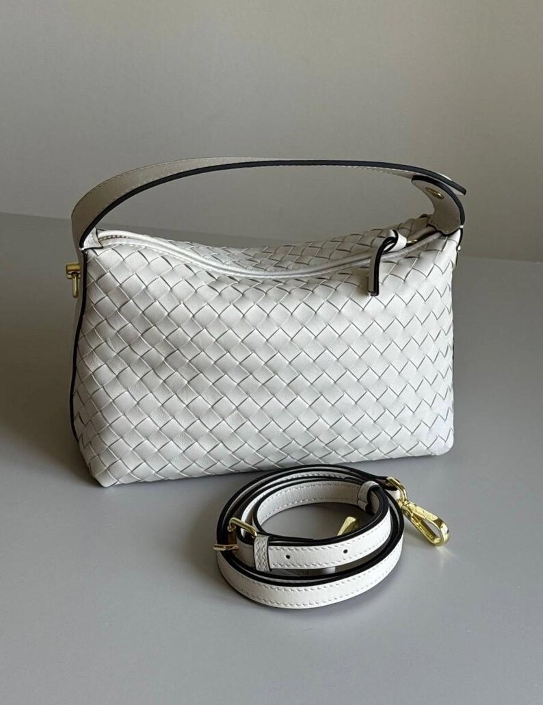 May include: White woven leather handbag with a top handle and a detachable shoulder strap. The bag features a zipper closure and gold-tone hardware. The strap is white with black trim and gold-tone clasps.