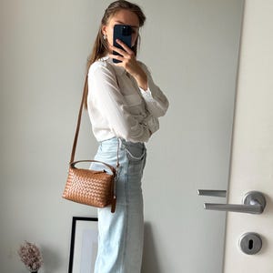 May include: A brown woven leather crossbody bag with a top handle and a long adjustable strap. The bag is rectangular with a woven pattern and a small leather tag. The bag is worn over the shoulder.