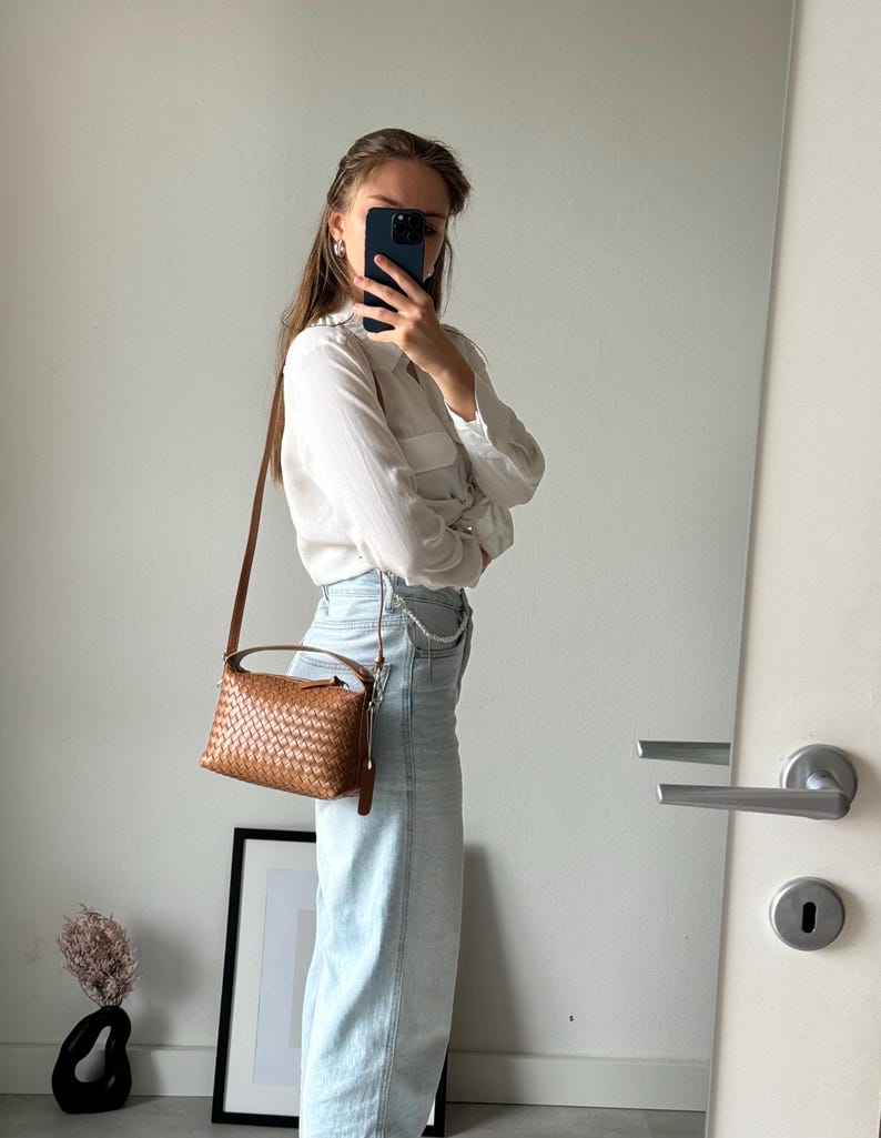 May include: A brown woven leather crossbody bag with a top handle and a long adjustable strap. The bag is rectangular with a woven pattern and a small leather tag. The bag is worn over the shoulder.