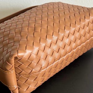 May include: A tan-colored handbag with a woven design. The bag features a rectangular shape with a shoulder strap. The woven pattern creates a textured surface, and the bag appears to be made of leather.