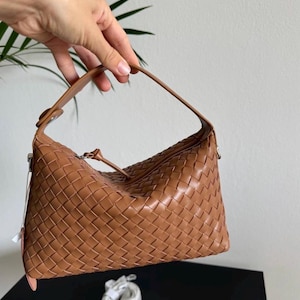 May include: A woven leather handbag in a warm brown hue. The bag features a distinctive basketweave pattern and a single handle. The bag is held up by a hand, showcasing its shape and design. A small, white accessory is visible below the bag.