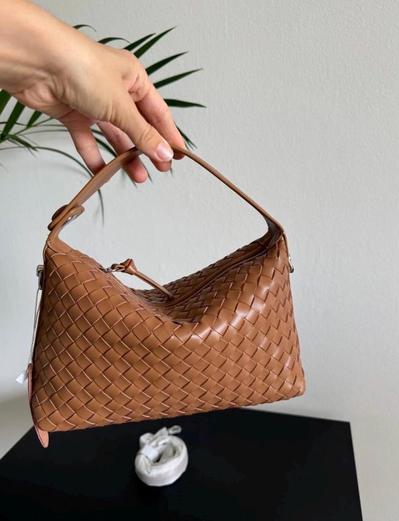 May include: A woven leather handbag in a warm brown hue. The bag features a distinctive basketweave pattern and a single handle. The bag is held up by a hand, showcasing its shape and design. A small, white accessory is visible below the bag.
