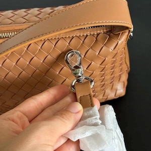 May include: A tan woven leather handbag with a silver-tone clasp and a matching leather strap. The bag has a zipper closure and a woven pattern. The strap is attached to the bag with a silver-tone clasp.