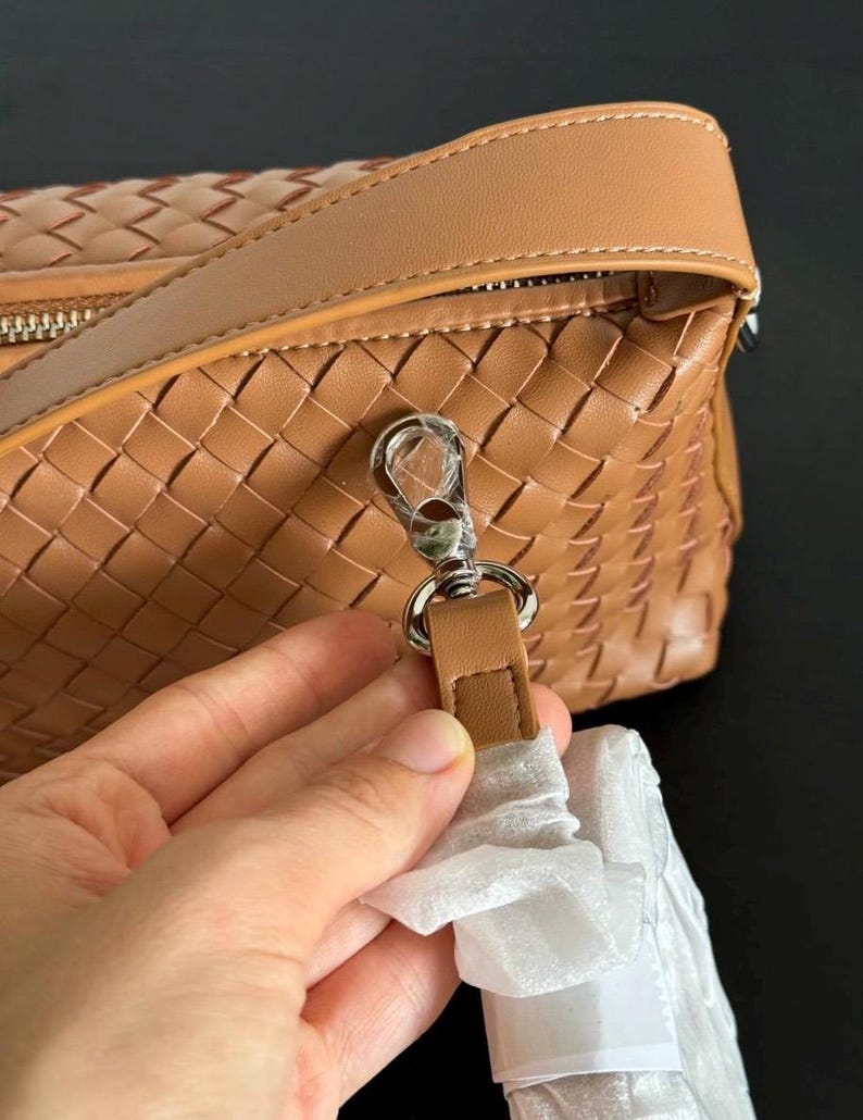 May include: A tan woven leather handbag with a silver-tone clasp and a matching leather strap. The bag has a zipper closure and a woven pattern. The strap is attached to the bag with a silver-tone clasp.