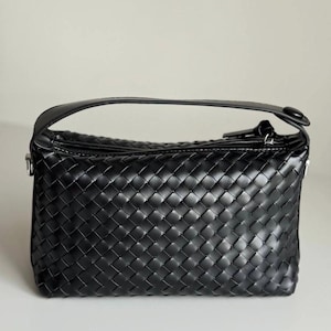 May include: Black woven leather handbag with a top handle. The bag features a rectangular shape with a zipper closure and a woven pattern. The handle is a solid black leather. The bag is sitting on a white surface.