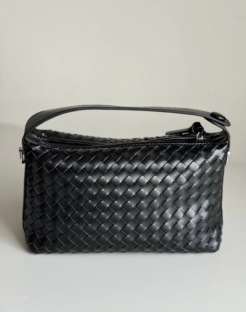 May include: Black woven leather handbag with a top handle. The bag features a rectangular shape with a zipper closure and a woven pattern. The handle is a solid black leather. The bag is sitting on a white surface.