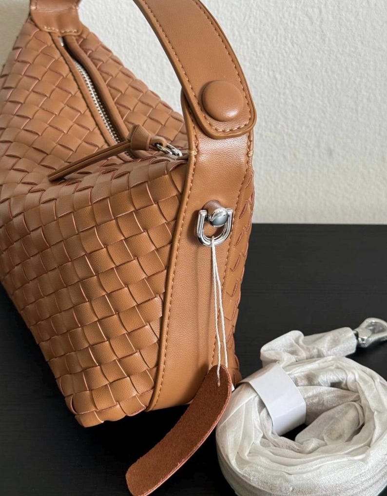 May include: A woven, tan-colored handbag with a rounded shape. The bag features a top handle with a button closure and a silver-toned metal clasp. A detachable strap and a leather tag are also included.