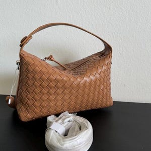 May include: A tan leather handbag with a woven design. The bag has a curved handle and a detachable shoulder strap. The bag is sitting on a black surface. The bag is a light brown color.