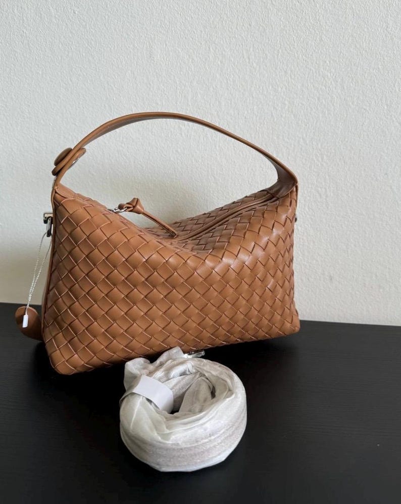 May include: A tan leather handbag with a woven design. The bag has a curved handle and a detachable shoulder strap. The bag is sitting on a black surface. The bag is a light brown color.