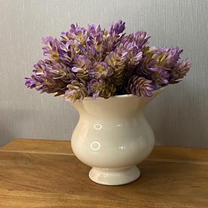 Pode incluir: Um vaso de cerâmica branco-sujo brilhante com uma borda larga e alargada e um corpo arredondado, preenchido com um arranjo denso de flores artificiais cor de lavanda com centros tingidos de verde e folhagem em camadas pelo buquê.