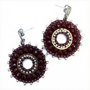Könnte beinhalten: Ein Paar handgefertigte Ohrringe mit kreisförmigem Design. Die Ohrringe haben einen tiefroten, perlenbesetzten Außenring, einen silber-goldenen Innenring und silberne Akzente. Die Ohrringe haben einen Steckerverschluss und sind vor weißem Hintergrund.