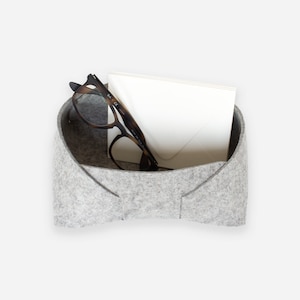 May include: A gray felt storage organizer with a folded collar shape. It contains a pair of eyeglasses and a white envelope.