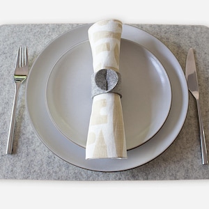 May include: A place setting with a light gray felt placemat, two white plates, a folded napkin with a gray napkin ring, a fork, and a knife. The napkin is beige with a white pattern.