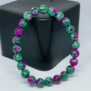May include: A bracelet made of round beads in shades of green, magenta, and black. The beads are strung together to form a circular shape. The bracelet is set against a white backdrop with a black square.