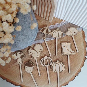 Puede incluir: Una colección de palillos decorativos de madera con temática otoñal. El conjunto incluye un paraguas, calabazas, un erizo, una seta, una ardilla, una bellota y botas de agua. Los palillos están dispuestos sobre una rodaja de madera.